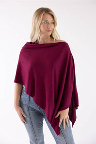 A woman modeling a deep burgundy off-the-shoulder poncho top paired with light-wash blue jeans against a plain white studio background.
