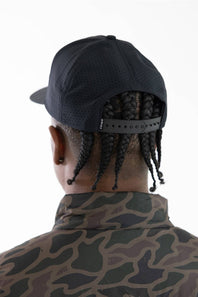 Rear view of a man wearing a black perforated baseball cap with an adjustable strap and a camouflage-patterned jacket, against a plain white background.