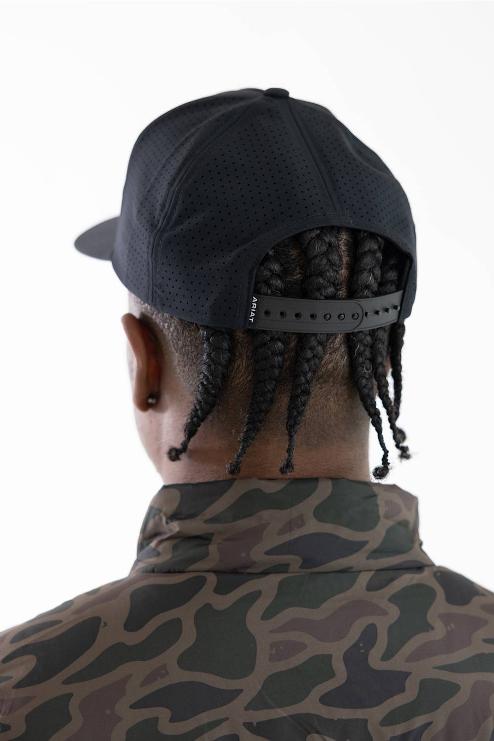 Rear view of a man wearing a black perforated baseball cap with an adjustable strap and a camouflage-patterned jacket, against a plain white background.