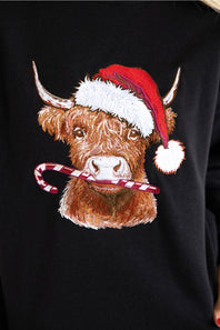 A close-up of a black sweatshirt featuring a detailed embroidered graphic of a brown highland cow wearing a red Santa hat and holding a candy cane.