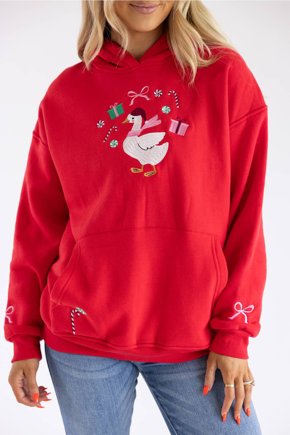 A woman wearing a bright red pullover hoodie with a white goose and holiday-themed embroidery on the chest, paired with blue jeans against a plain white background.