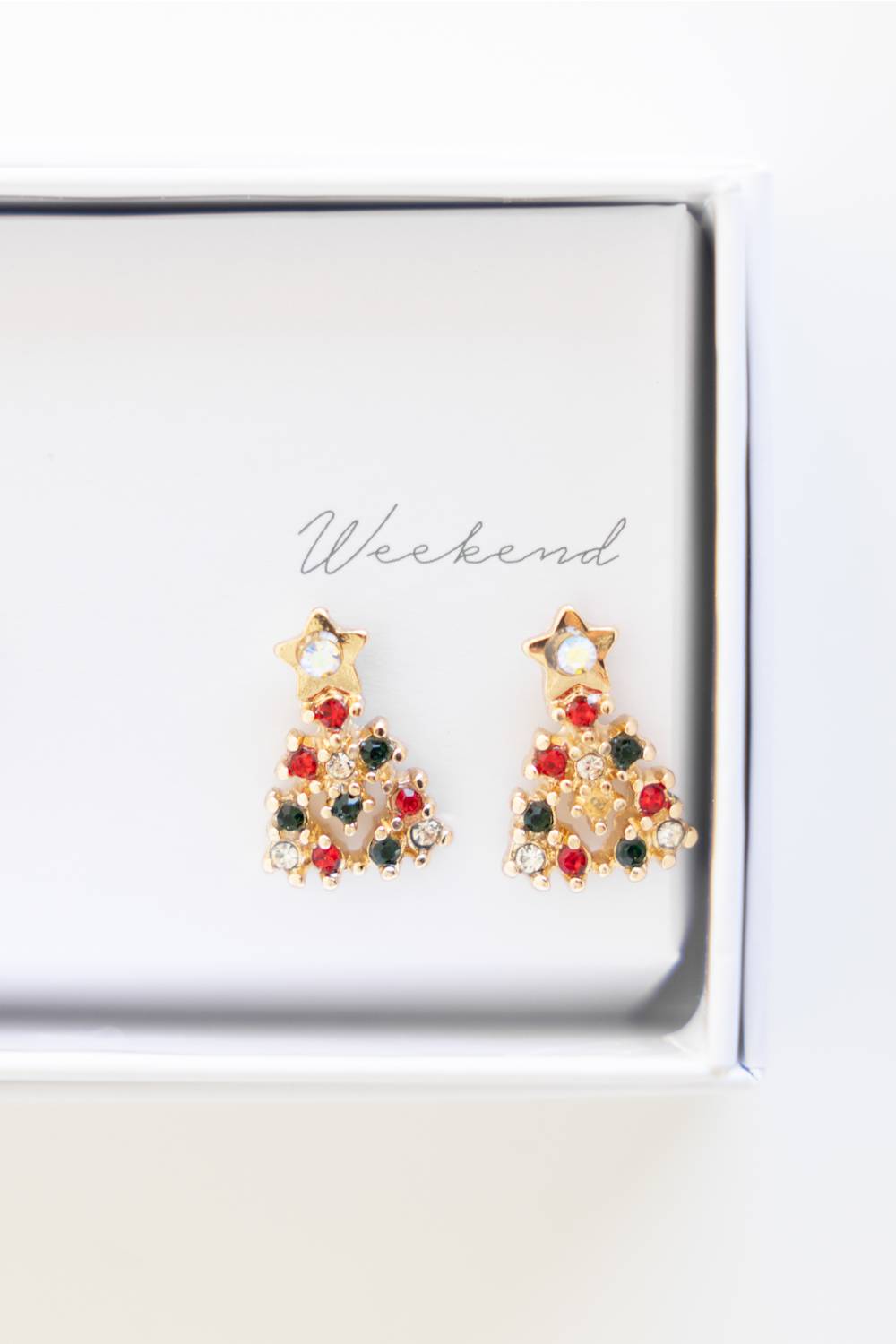 A pair of gold-toned dangle earrings shaped like Christmas trees, decorated with red, green, and clear rhinestones, resting on a white jewelry box insert with the brand name Weekend.