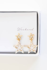 A pair of gold star-shaped dangle earrings with clear rhinestones, displayed inside a white jewelry box with the word 'Weekend' printed on the inside lid.