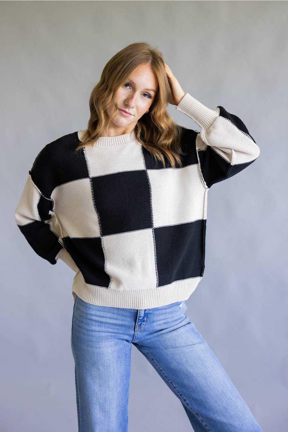 A woman wearing a black and off-white checkered knit sweater with blue jeans, posing against a neutral gray studio background.
