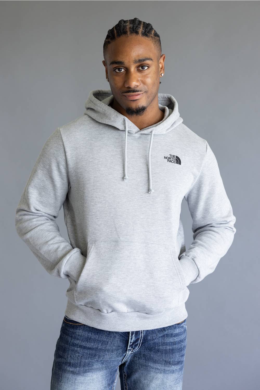 A man wearing a light gray heathered hoodie with a small black embroidered logo on the left chest, paired with blue jeans, standing against a solid gray studio background.