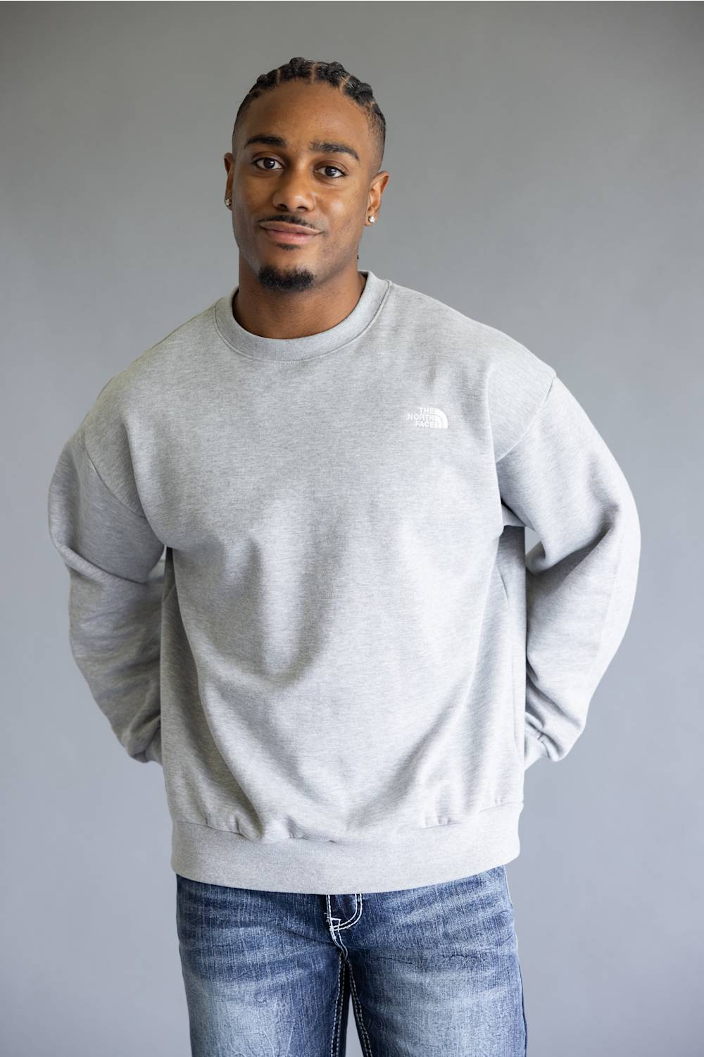 A man wearing a light gray heather crewneck sweatshirt by The North Face, paired with blue jeans, standing against a solid gray studio background.