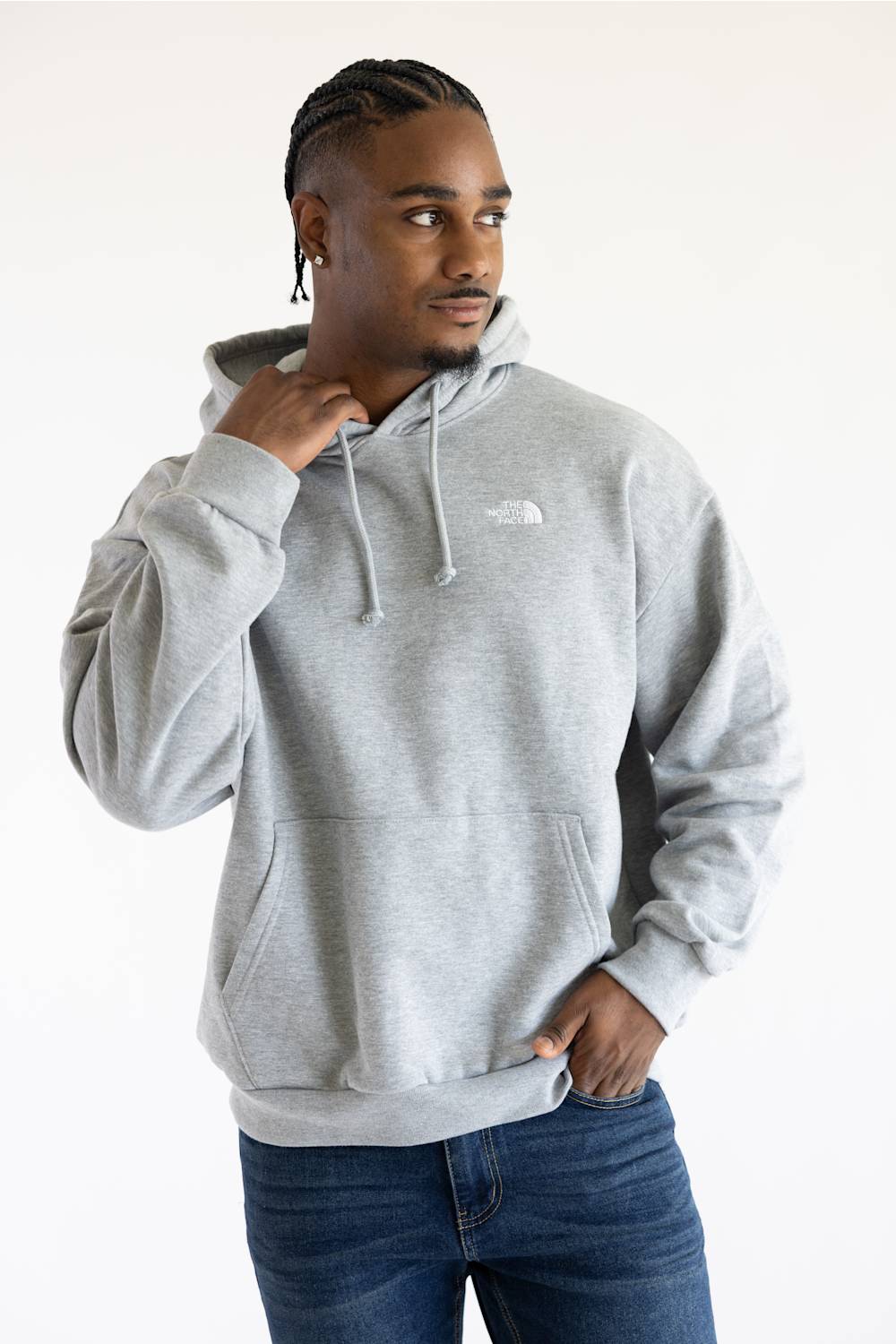 A man wearing a light gray heather pullover hoodie with a small white embroidered logo on the chest, paired with dark blue jeans, photographed against a plain white studio background.