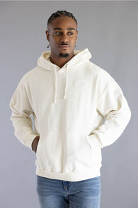 A man wearing an oversized off-white pullover hoodie with drawstrings and a kangaroo pocket, paired with blue jeans, standing against a solid gray studio background.