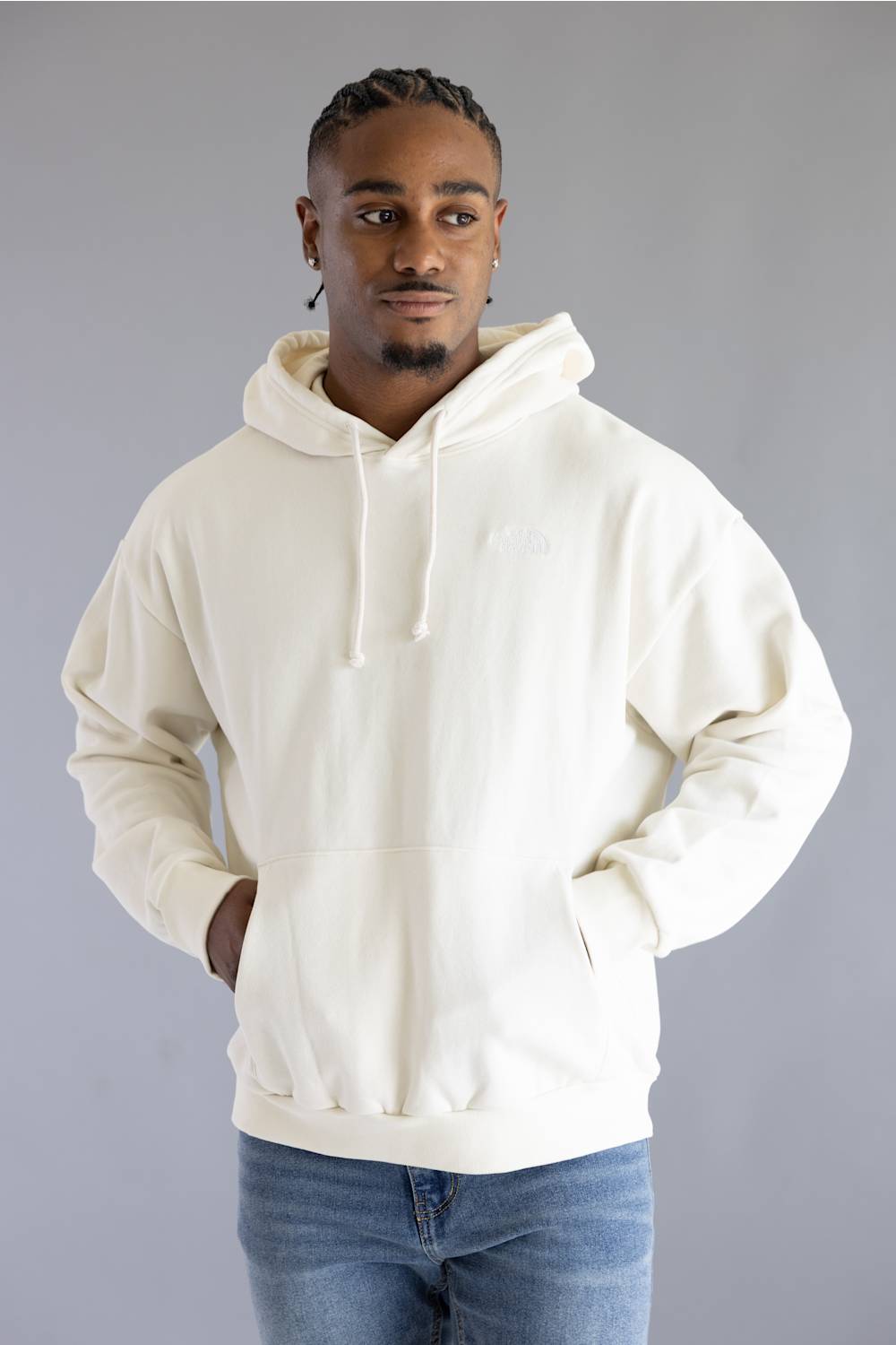 A man wearing an oversized off-white pullover hoodie with drawstrings and a kangaroo pocket, paired with blue jeans, standing against a solid gray studio background.