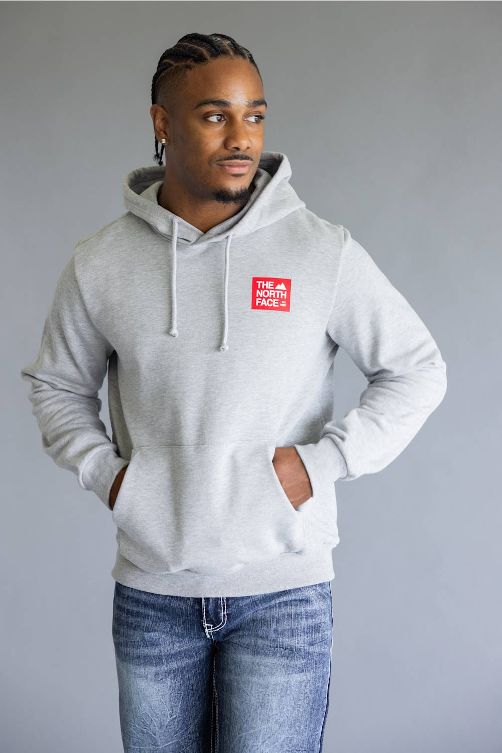 A man wearing a light gray heather pullover hoodie with a red 'The North Face' logo patch on the left chest, paired with blue jeans, against a solid gray studio background.