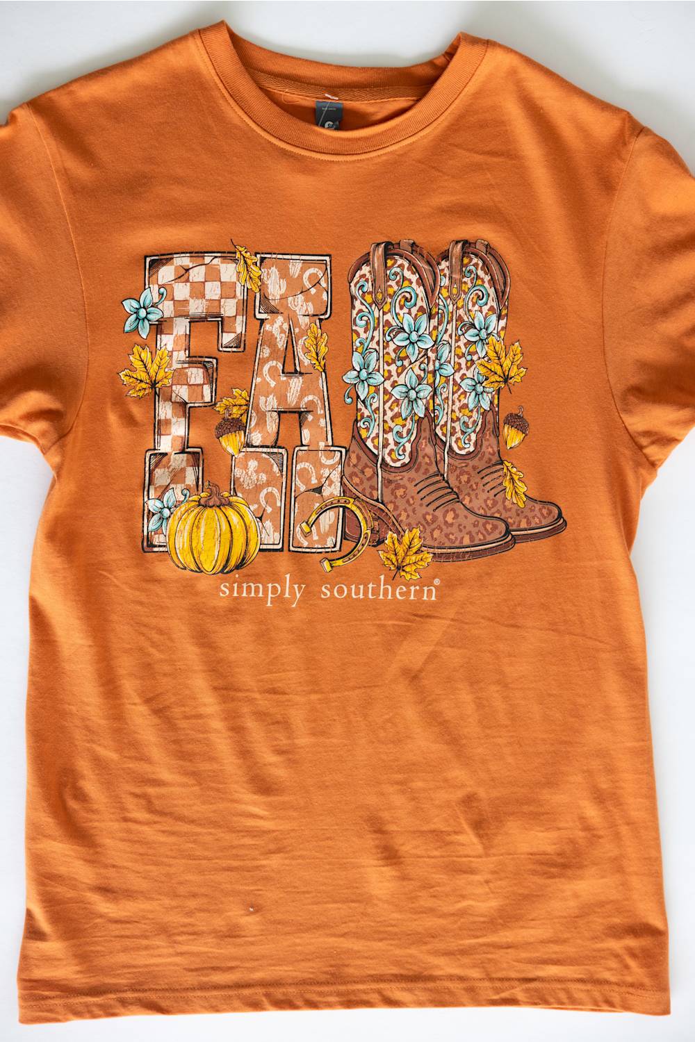 An orange short-sleeve crewneck t-shirt featuring a 'FALL' graphic with patterned letters, cowboy boots, pumpkins, and autumn leaves, with 'simply southern' printed below.