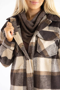 A close-up of a woman wearing a brown, beige, and gray plaid flannel shirt jacket with a matching brown cowl scarf, against a plain white background.