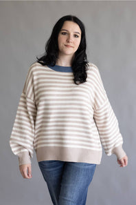 A woman modeling a cream and beige horizontally striped knit sweater with a contrasting blue crew neck, paired with blue jeans, against a plain gray studio background.