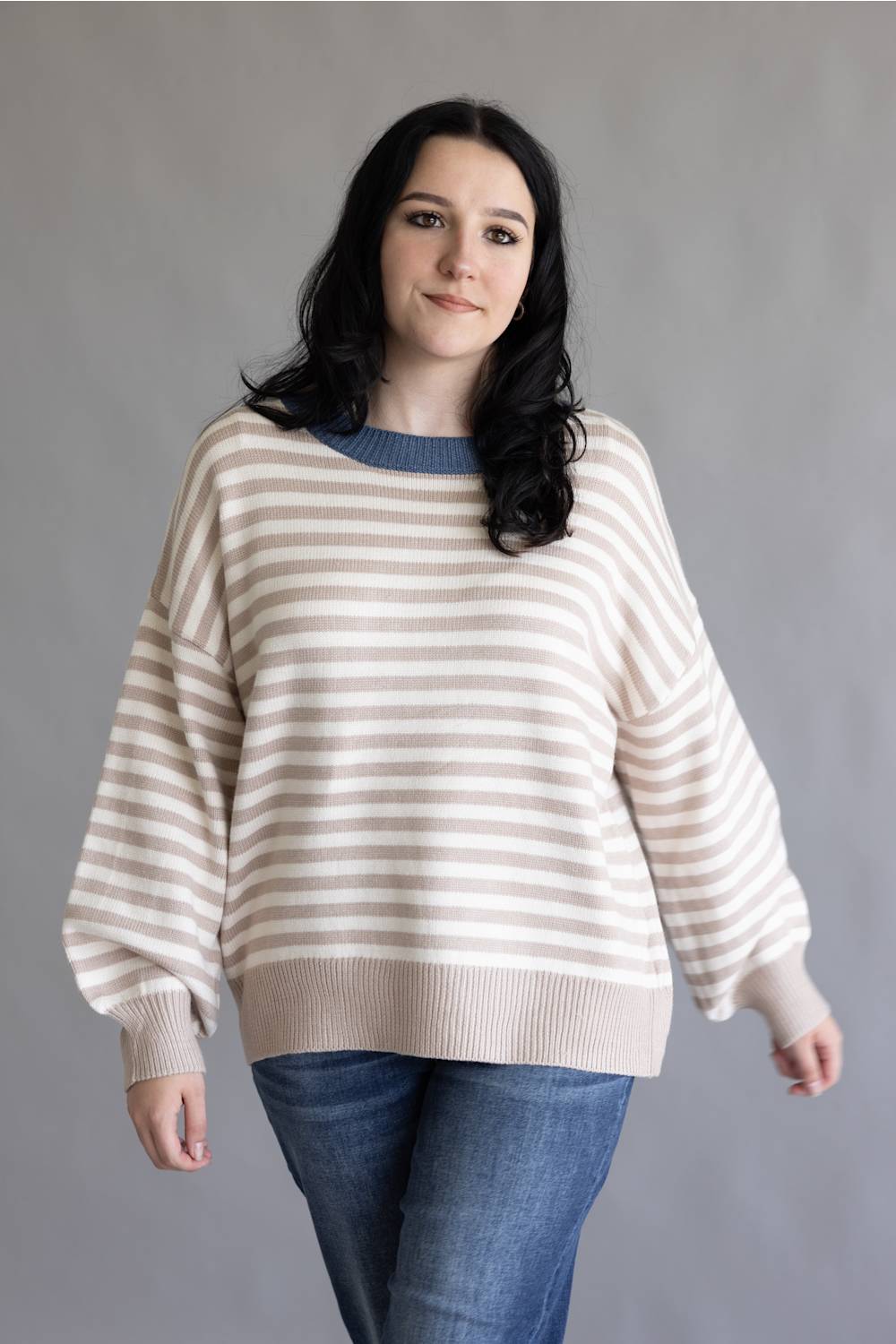 A woman modeling a cream and beige horizontally striped knit sweater with a contrasting blue crew neck, paired with blue jeans, against a plain gray studio background.