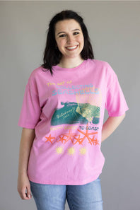 A woman wearing a pink graphic t-shirt with a coastal design featuring a green turtle, orange starfish, and yellow text, paired with blue jeans, against a plain gray background.