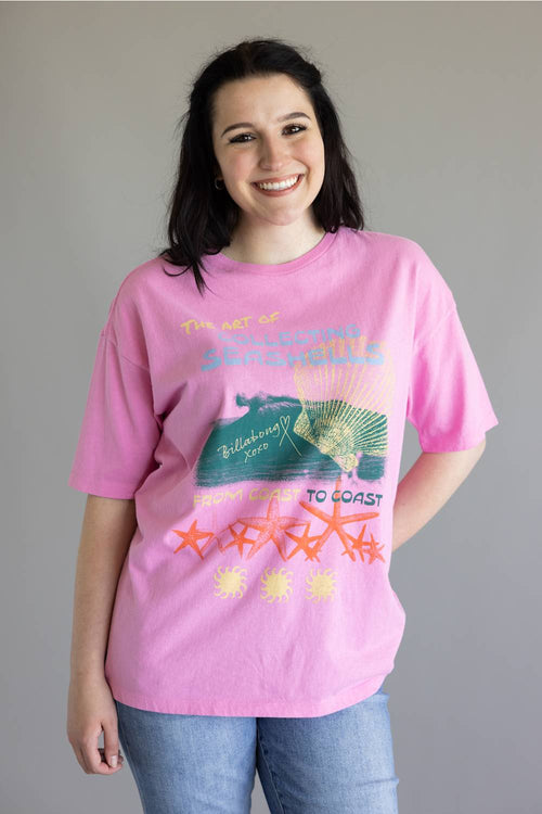 A woman wearing a pink graphic t-shirt with a coastal design featuring a green turtle, orange starfish, and yellow text, paired with blue jeans, against a plain gray background.