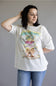 A woman wearing a white short-sleeve graphic t-shirt with a colorful vintage-style tropical print and blue jeans, standing against a plain gray studio background.