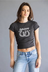 A woman modeling a dark charcoal gray crop top t-shirt with a white Billabong 1975 horseshoe graphic, paired with light wash blue jeans, against a solid gray studio background.