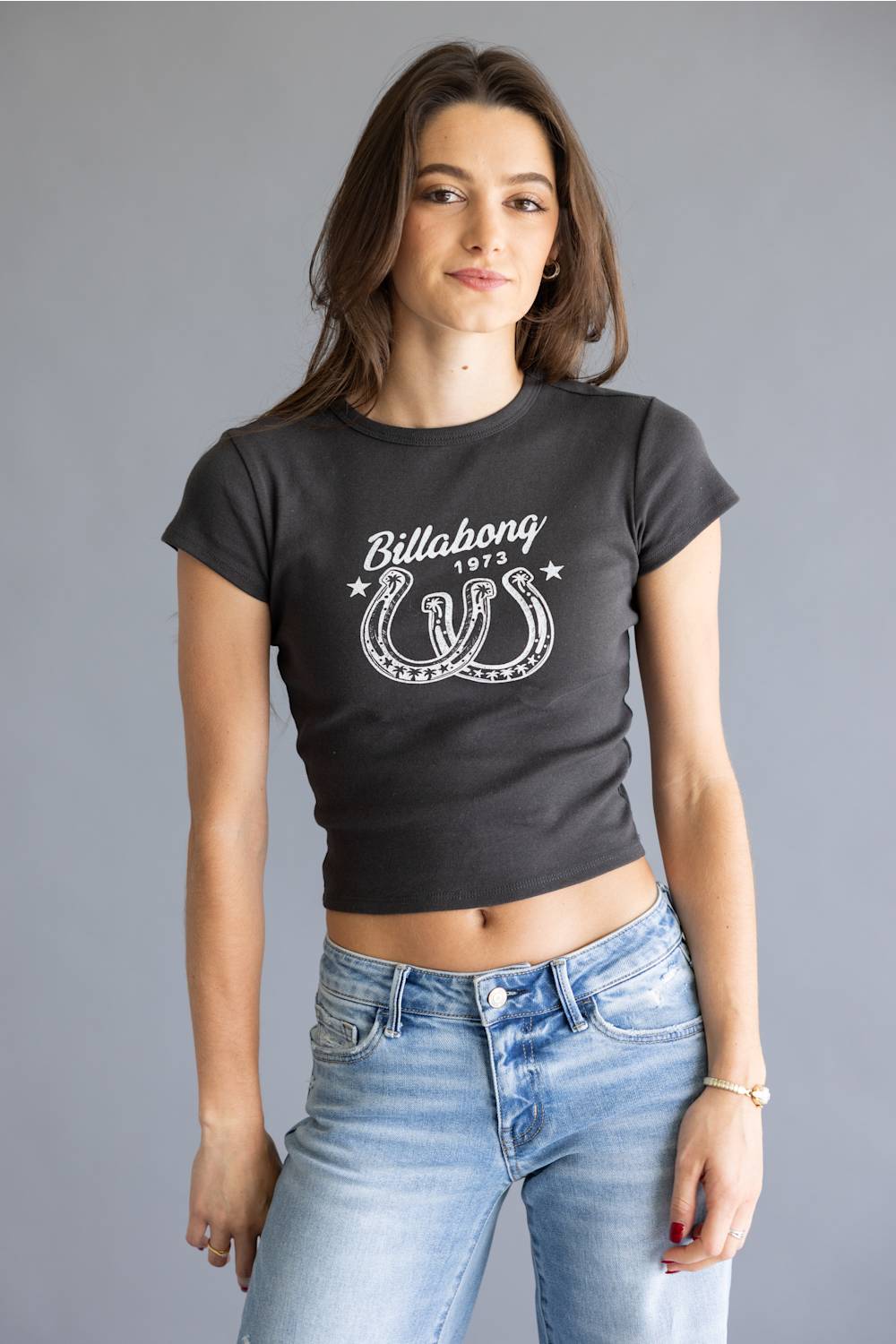 A woman modeling a dark charcoal gray crop top t-shirt with a white Billabong 1975 horseshoe graphic, paired with light wash blue jeans, against a solid gray studio background.