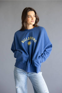 A woman wearing a blue Billabong sweatshirt with a yellow sun logo and text, paired with light-wash jeans, against a solid gray studio background.