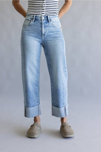A person wearing light-wash blue cropped jeans with rolled cuffs, paired with a black-and-white striped top and taupe slip-on shoes, standing against a light gray studio background.
