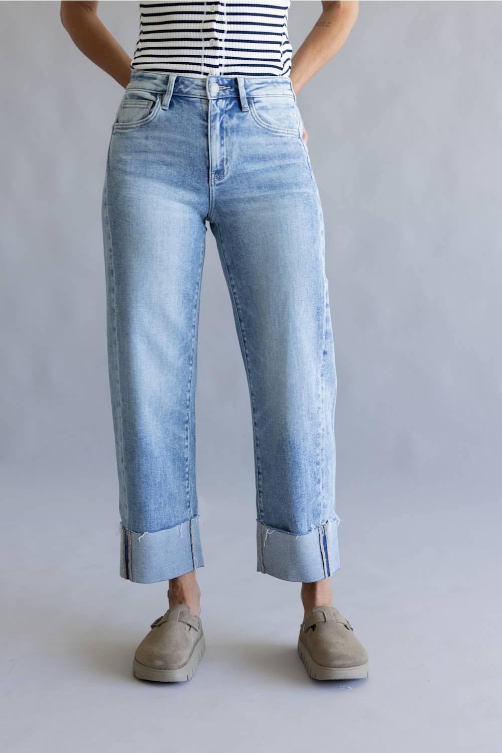 A person wearing light-wash blue cropped jeans with rolled cuffs, paired with a black-and-white striped top and taupe slip-on shoes, standing against a light gray studio background.