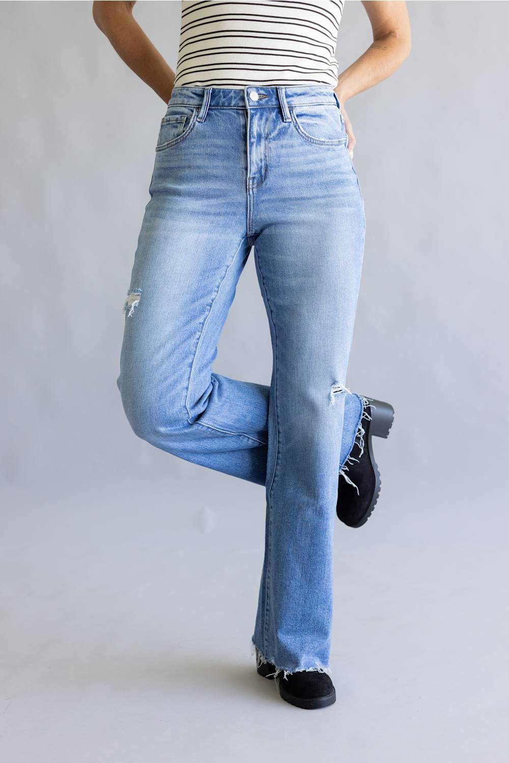 A person wearing light-wash blue high-rise flared jeans with distressed details on the knees, paired with a black and white striped top and black lace-up shoes, against a light gray studio background.