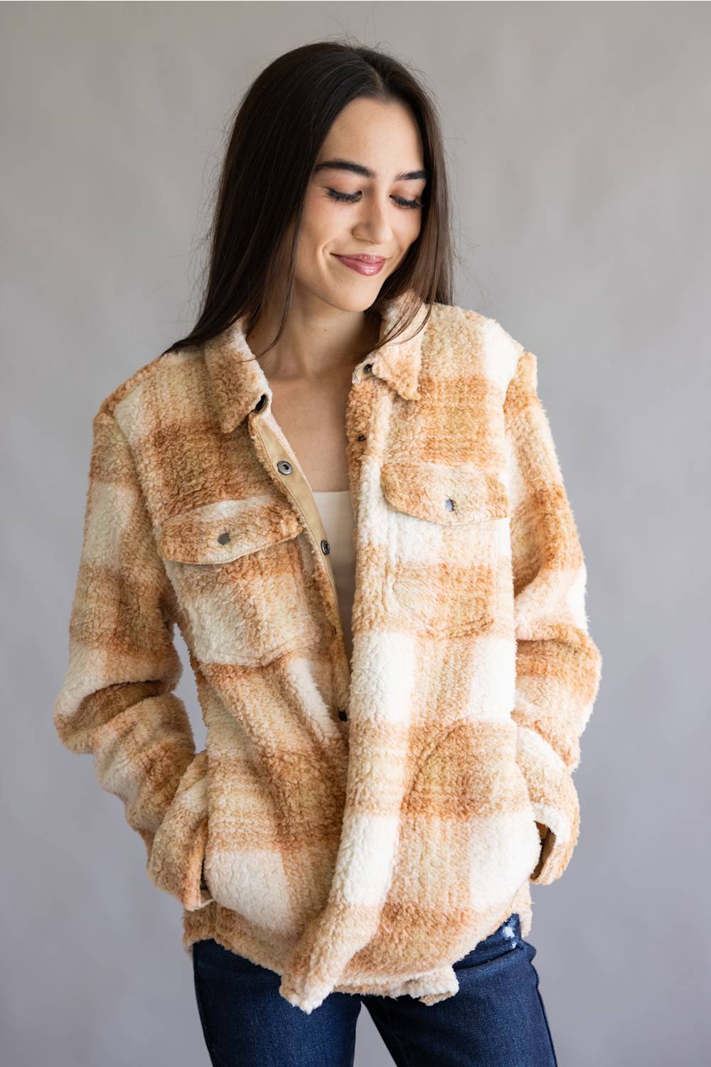 A woman wearing a fuzzy plaid button-up shirt jacket in beige, cream, and light brown tones, paired with a white top and dark blue jeans, set against a plain gray studio background.