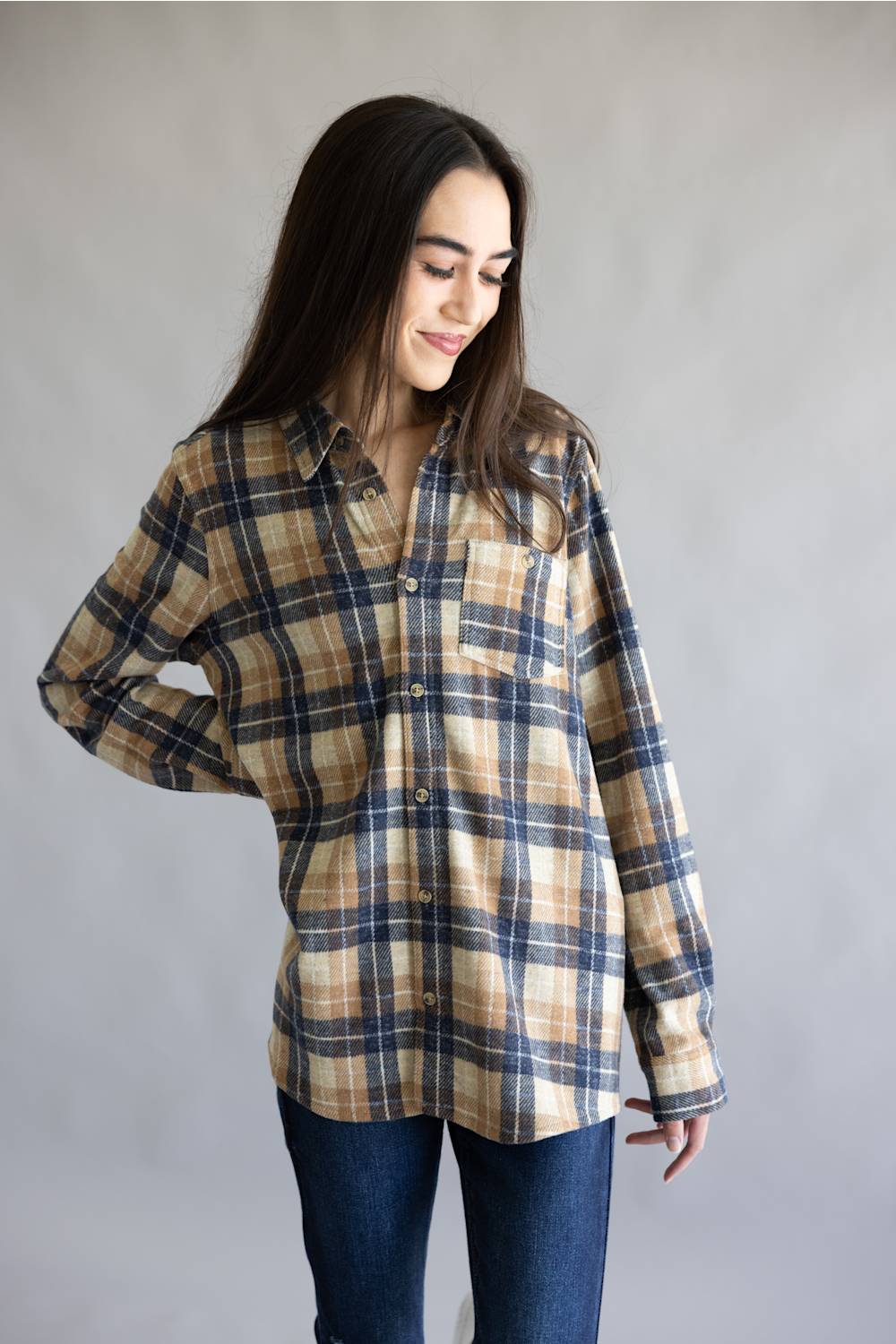 A woman wearing a beige, navy, and brown plaid button-up shirt with dark blue jeans, standing against a neutral gray studio background.