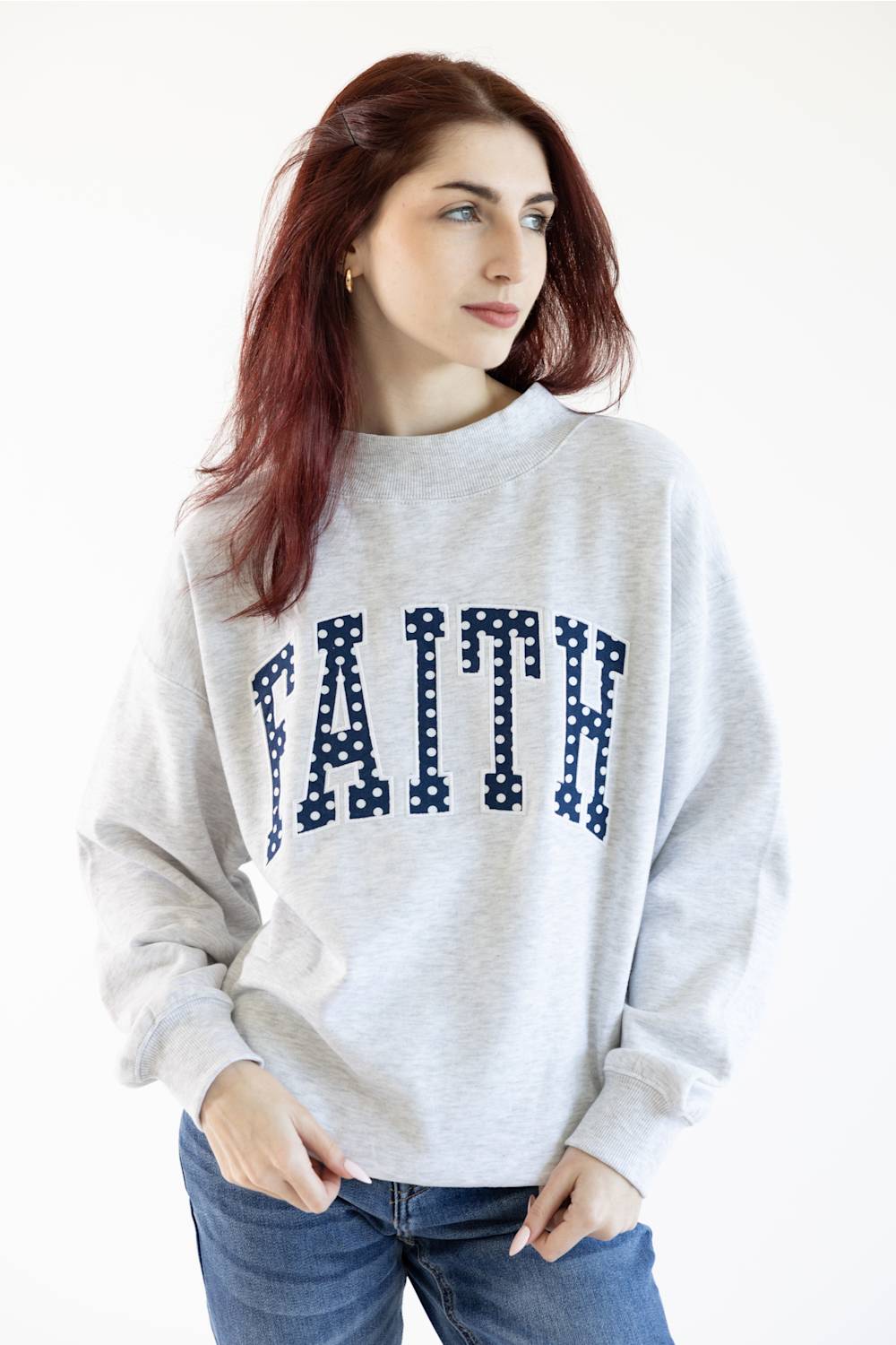 A woman with red hair wearing a light gray heather crewneck sweatshirt with the word 'FAITH' printed across the chest in navy blue block letters filled with white polka dots, paired with blue jeans.