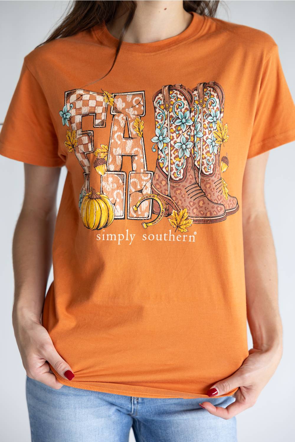 A woman wearing a bright orange graphic t-shirt with a 'FALL' design featuring cowboy boots, pumpkins, and flowers, paired with blue jeans.
