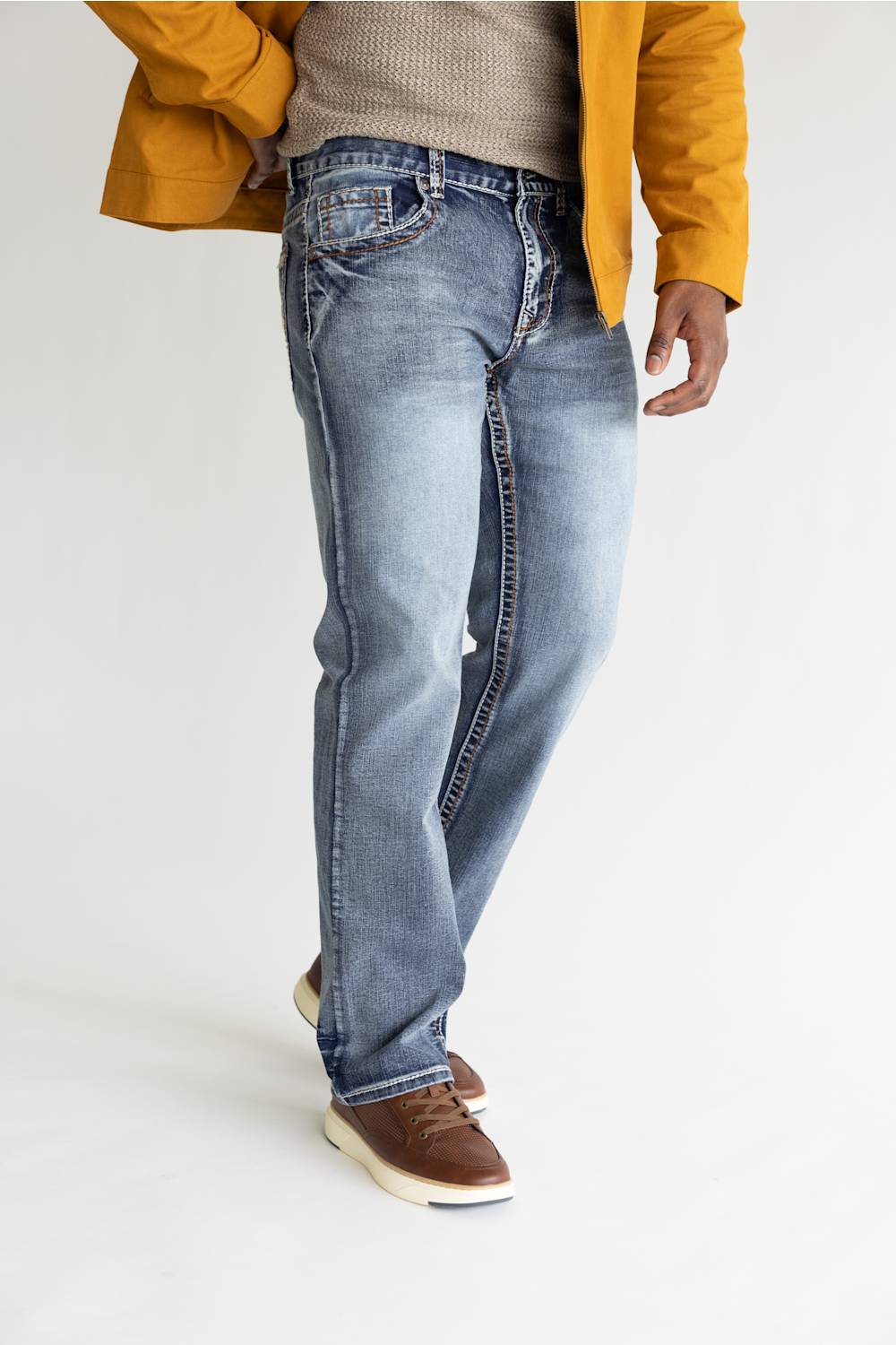 A person wearing light-wash blue jeans, a beige knit sweater, a mustard yellow jacket, and brown leather shoes, standing against a plain white studio background.