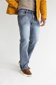 A person wearing light-wash blue jeans, a beige knit sweater, a mustard yellow jacket, and brown leather shoes, standing against a plain white studio background.