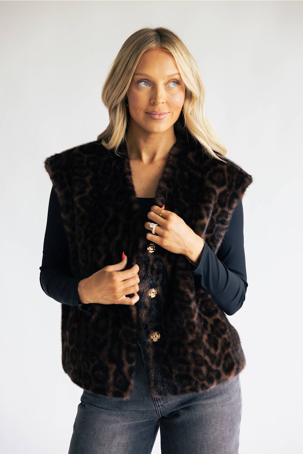 A blonde woman wearing a dark brown and black leopard-print faux fur vest over a black long-sleeve top and gray jeans, standing against a light gray studio background.