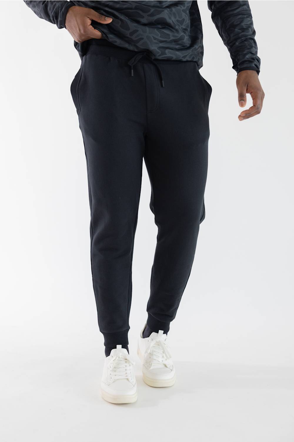 A person wearing jogger-style pants with a drawstring waist, paired with white sneakers and a dark patterned top, photographed against a clean white studio background.