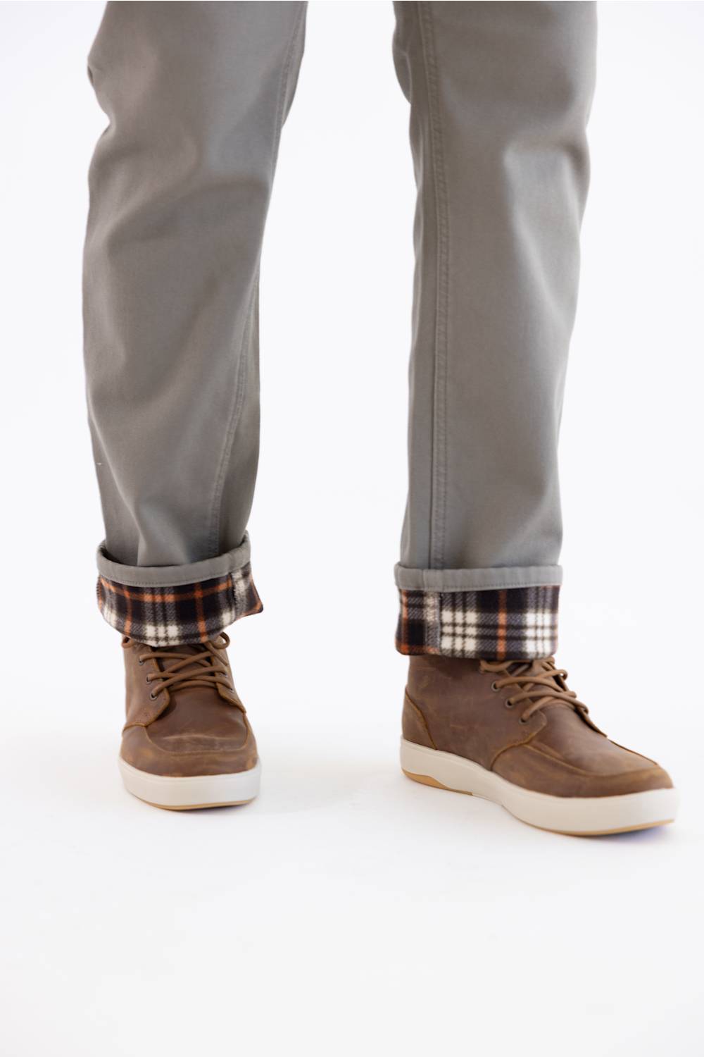 A close-up of a person's legs from the knees down, wearing gray pants with a plaid lining visible at the cuffs, paired with brown suede lace-up shoes on a white background.