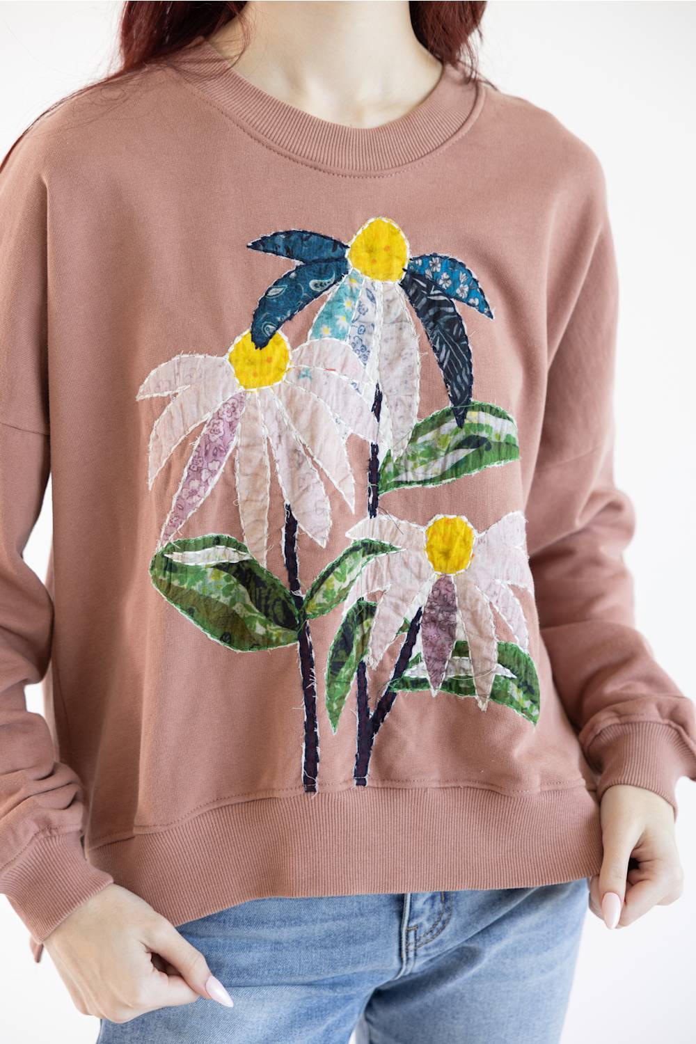 A person wearing a dusty rose crewneck sweatshirt with a large, colorful patchwork floral applique on the front, paired with blue jeans against a plain white background.
