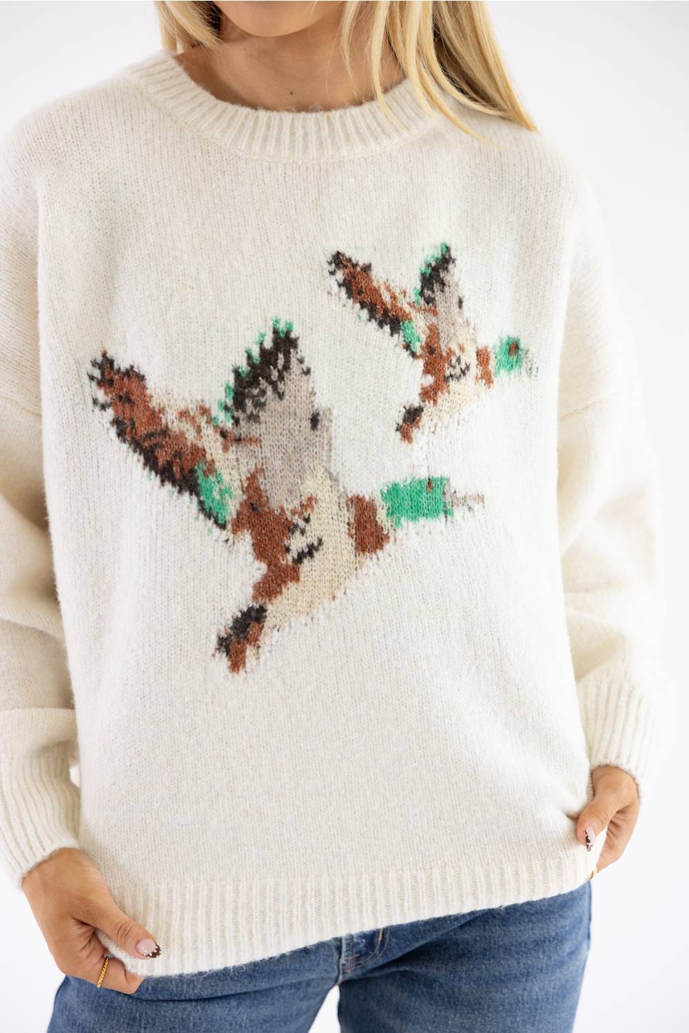 A close-up of a person wearing a cream-colored knit sweater with a large, textured bird motif in brown, beige, and green across the chest, paired with blue jeans.