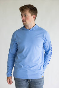 A man wearing a light blue long-sleeve hooded shirt with a vertical logo on the right sleeve, paired with blue jeans, standing against a plain light gray background.