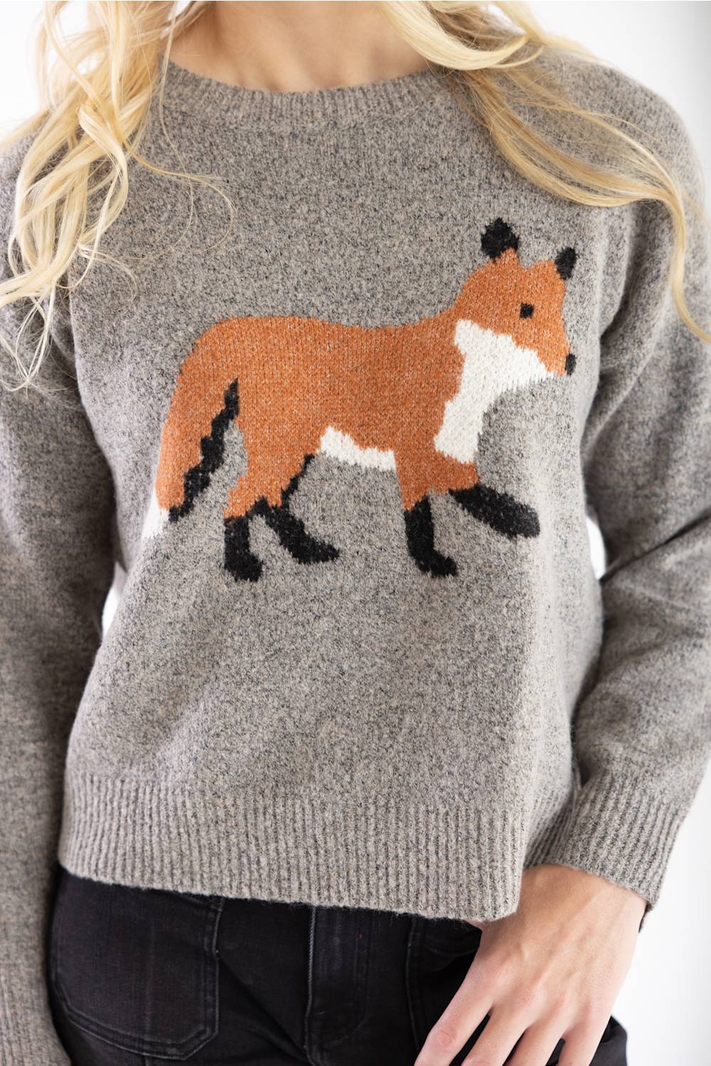 A woman wearing a heather gray knit sweater with a large knitted fox design on the front in orange, white, and black, paired with black jeans.
