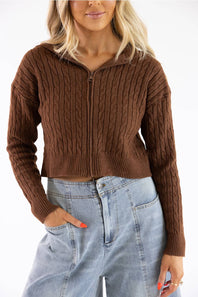 A woman wearing a brown cable-knit zip-up sweater with a mock neck and light-wash blue jeans, shown from the waist up against a plain white background.