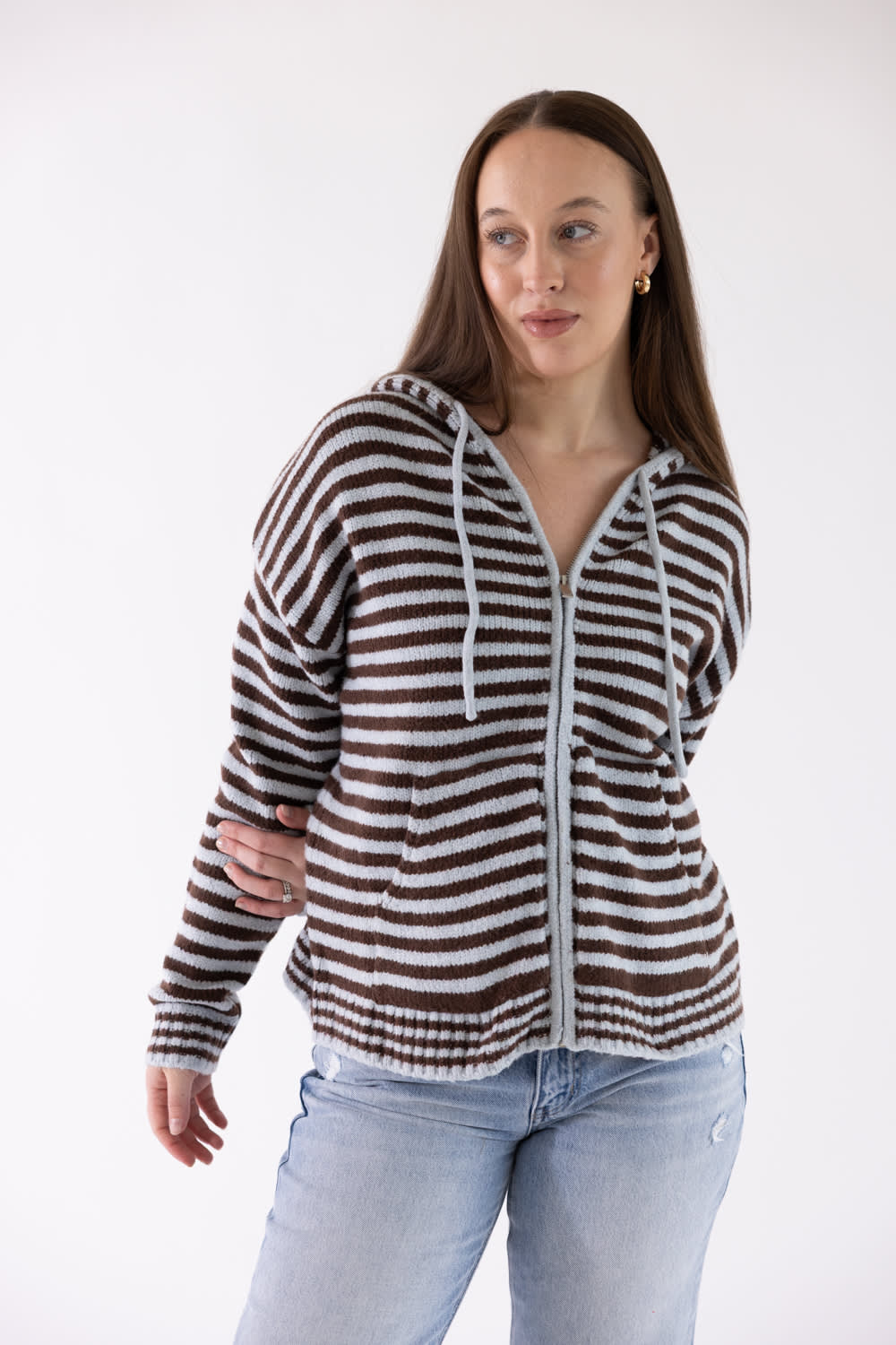 A woman modeling a brown and white horizontally striped zip-up hoodie with white drawstrings, paired with light-wash blue jeans, against a plain white studio background.