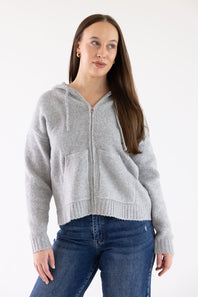 A woman wearing a light gray zip-up hooded sweater with drawstrings and front pockets, paired with blue jeans, photographed against a plain white studio background.