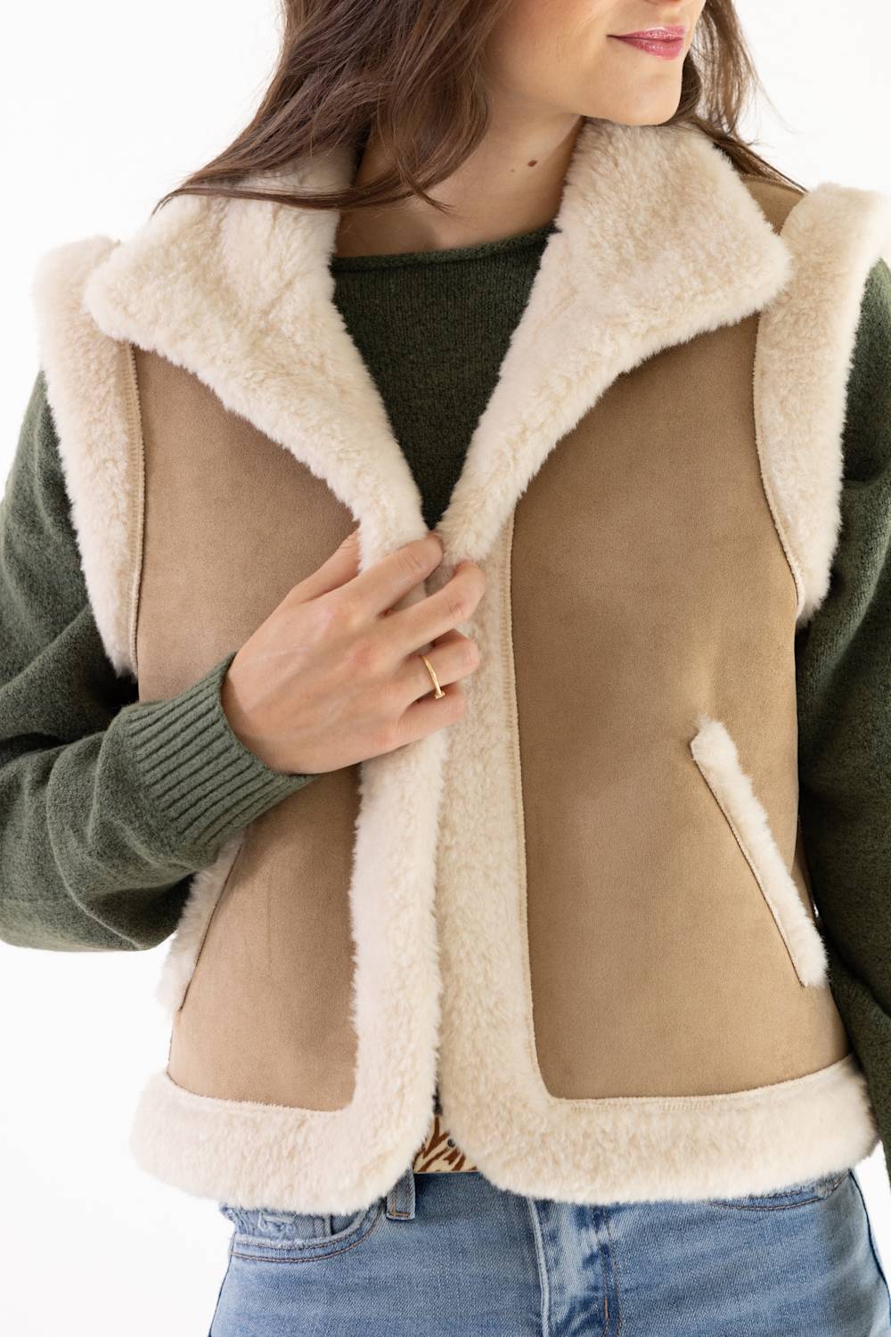A woman wearing a tan shearling-lined sleeveless vest over a dark green sweater and blue jeans, shown from the chest down against a plain white background.
