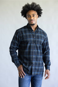 A man with an afro hairstyle wearing a dark blue and black plaid button-down shirt and blue jeans, standing against a plain light gray background.