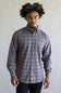 A man with an afro hairstyle wearing a long-sleeve button-down plaid shirt in red, blue, and gray, paired with black pants, standing against a plain light gray background.