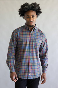 A man with an afro hairstyle wearing a long-sleeve button-down plaid shirt in red, blue, and gray, paired with black pants, standing against a plain light gray background.