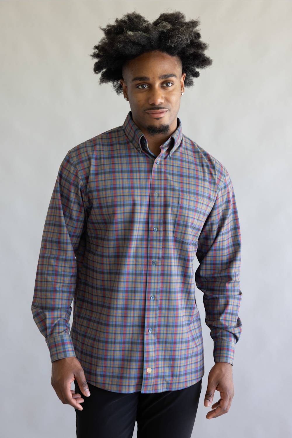 A man with an afro hairstyle wearing a long-sleeve button-down plaid shirt in red, blue, and gray, paired with black pants, standing against a plain light gray background.