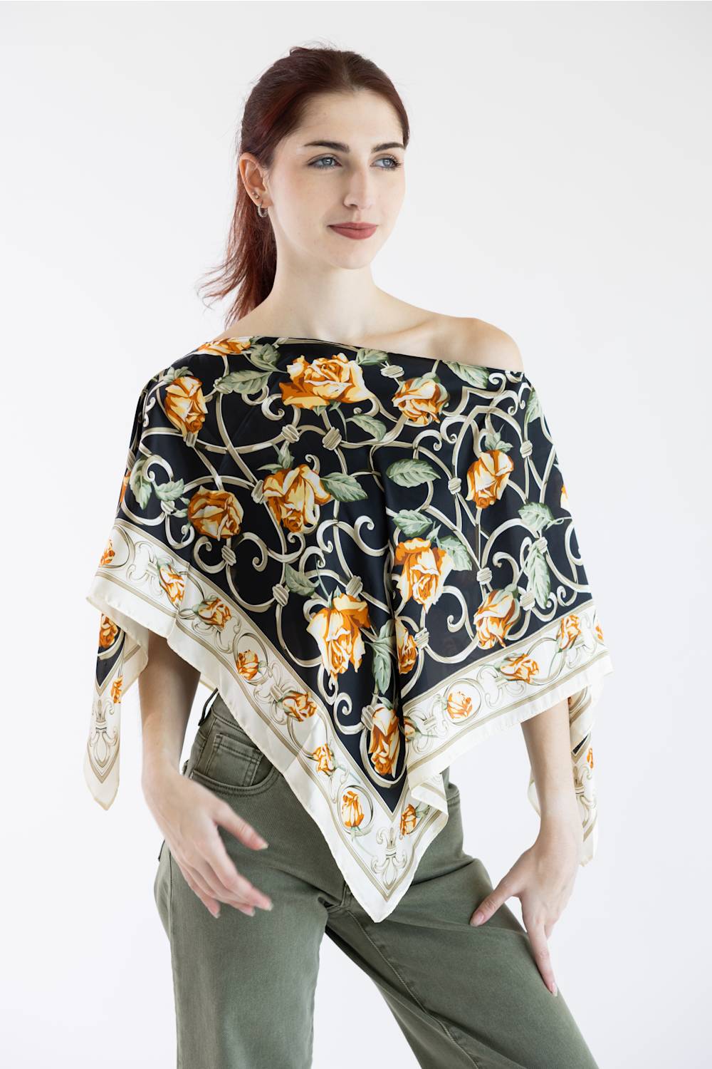 A woman modeling a square silk scarf draped off her shoulders. The scarf features a black background with a detailed print of orange roses, green leaves, and white scrolling vines, finished with a cream border.