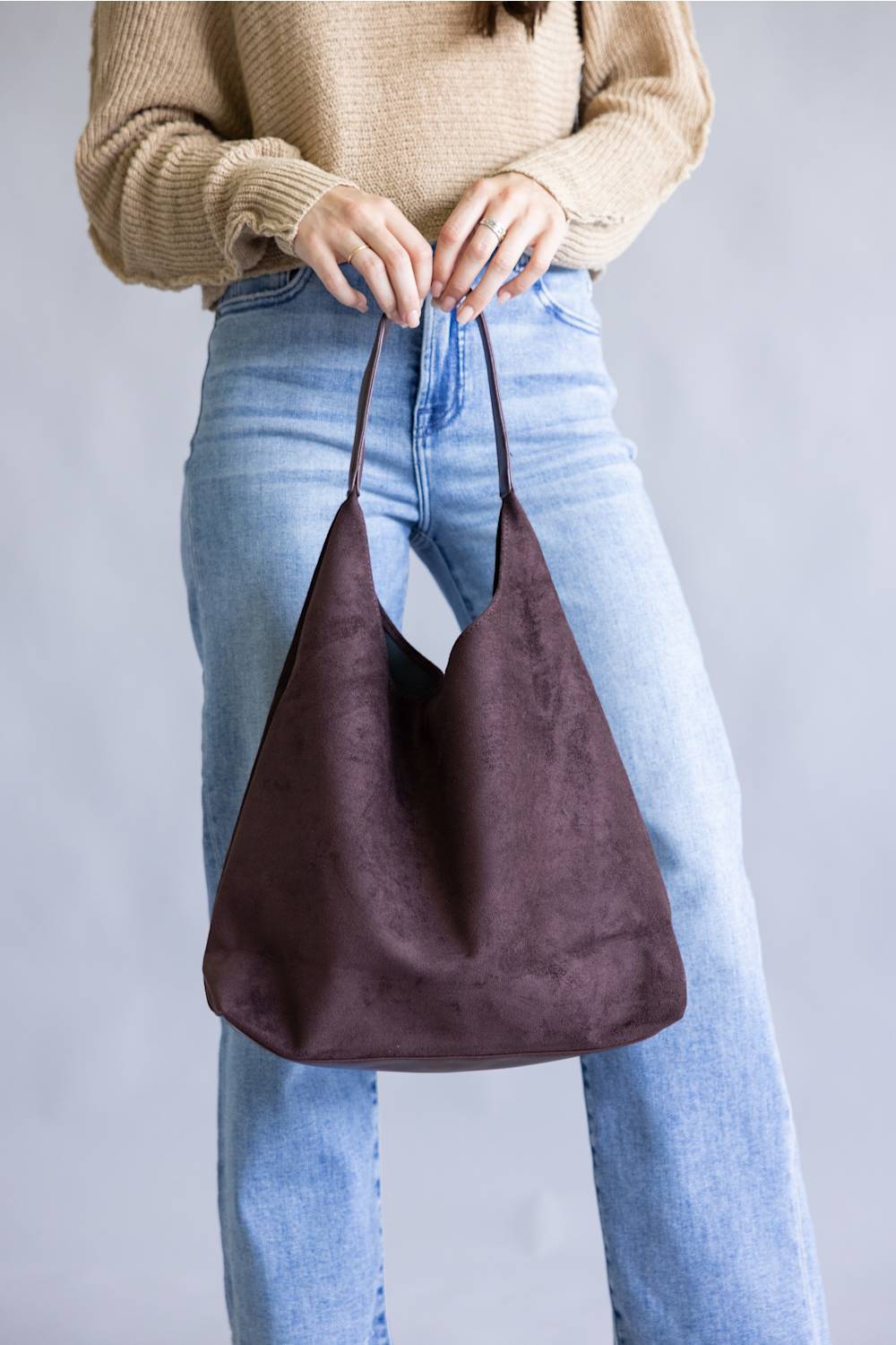 A person wearing a beige knit sweater and light blue jeans holds a large, slouchy hobo shoulder bag in dark brown suede against a plain gray studio background.